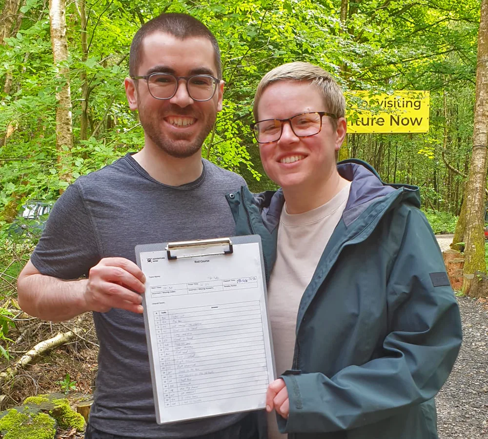 A smiling couple posing with their outdoor orienteering answer sheet