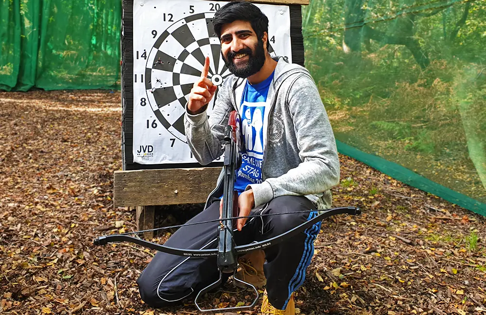 Smiling man crouched down, holding a crossbow in front of a crossbow target at Adventure Now Manchester