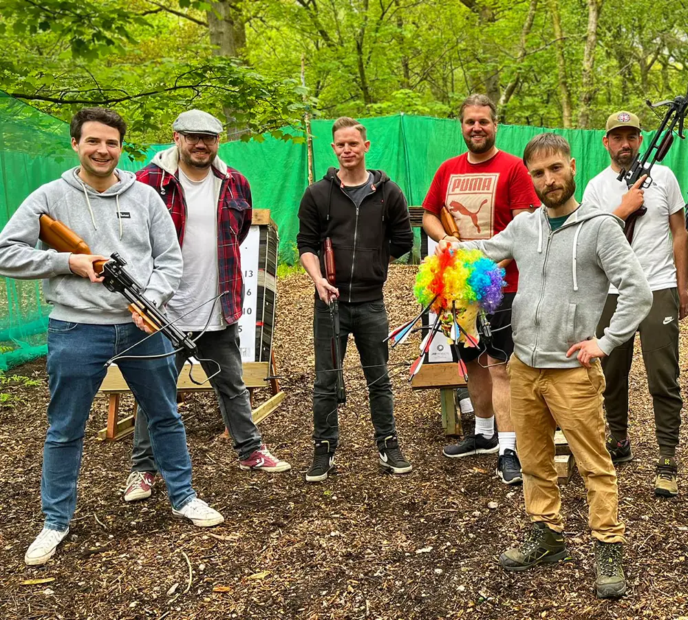 Group of 6 male friends smiling, holding crossbows and a zombie head with crossbow bolts stuck in it