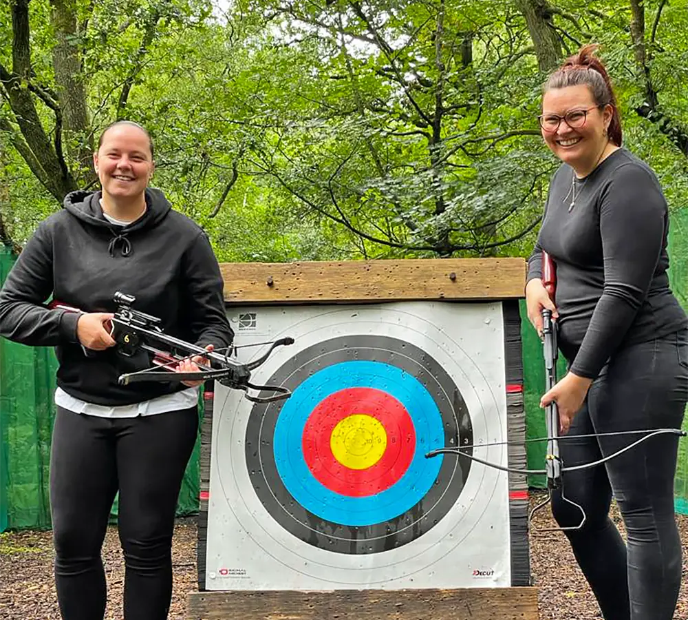 Two smiling women stood posing with crossbows and a crossbow target at Adventure Now