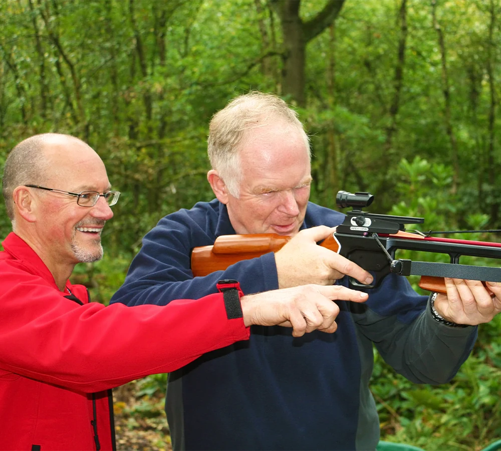 Smiling instructor pointing and participant smiling whilst aiming a wooden crossbow