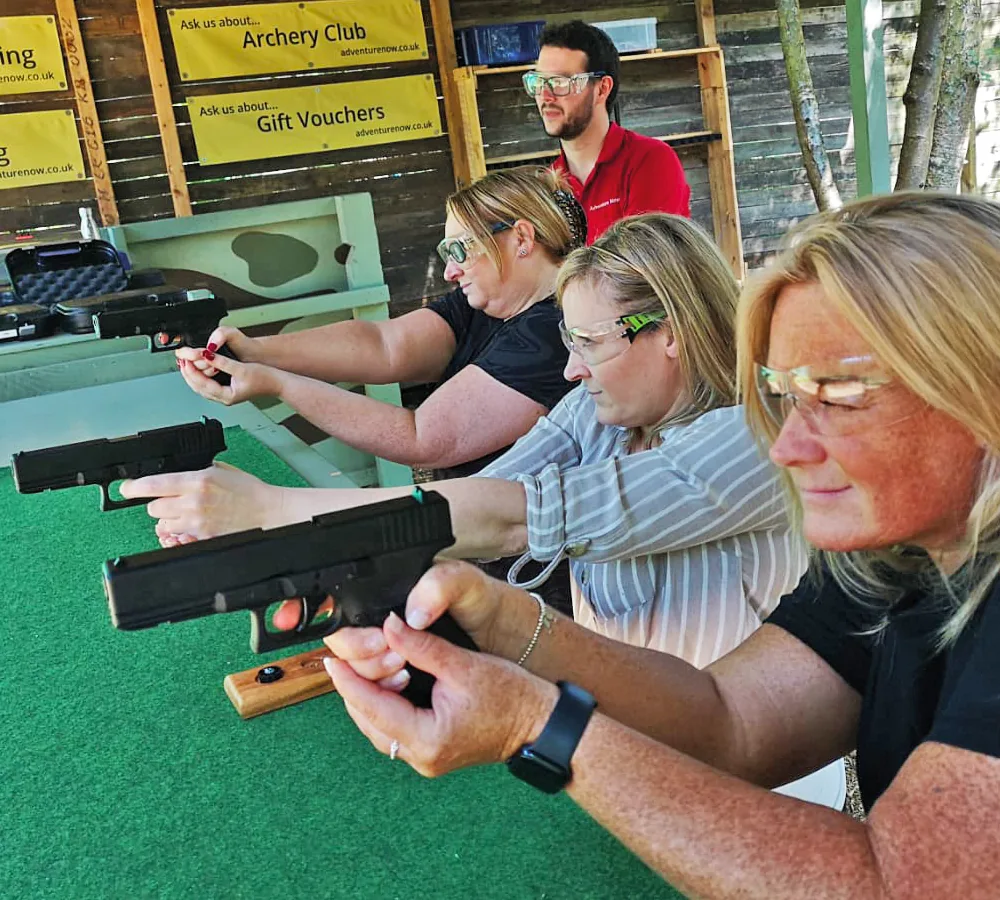 Three smiling women each aiming and shooting a Glock 17 pistol. Adventure Now instructor standing in the background