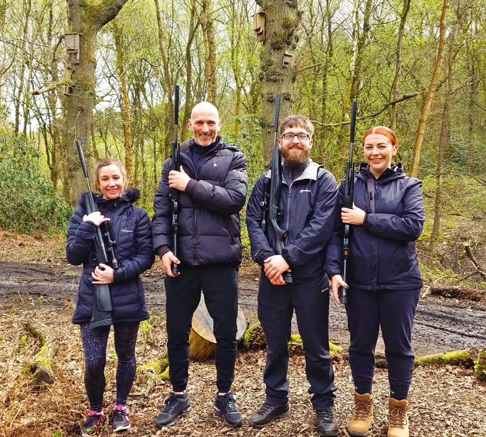 An adult family of four smiling and posing holding black Beretta air rifles at Adventure Now in Manchester