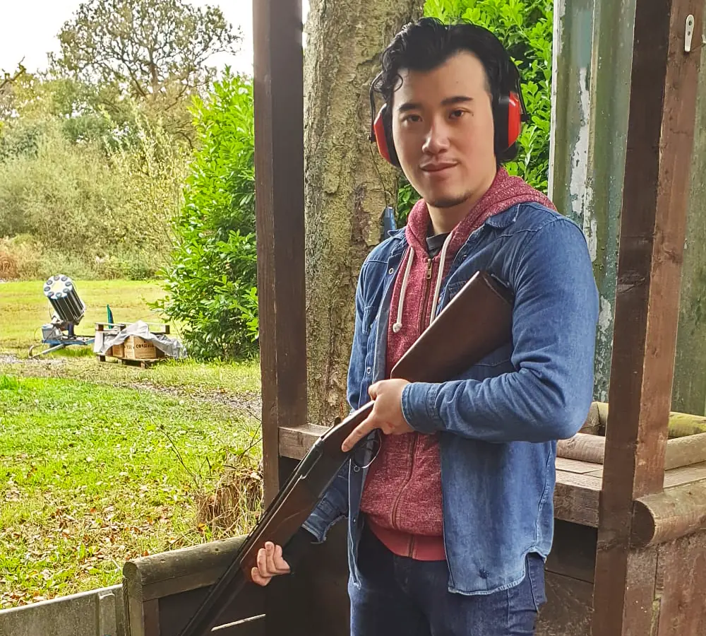 Smiling man posing with a shotgun wearing ear defenders, clay trap in background of picture