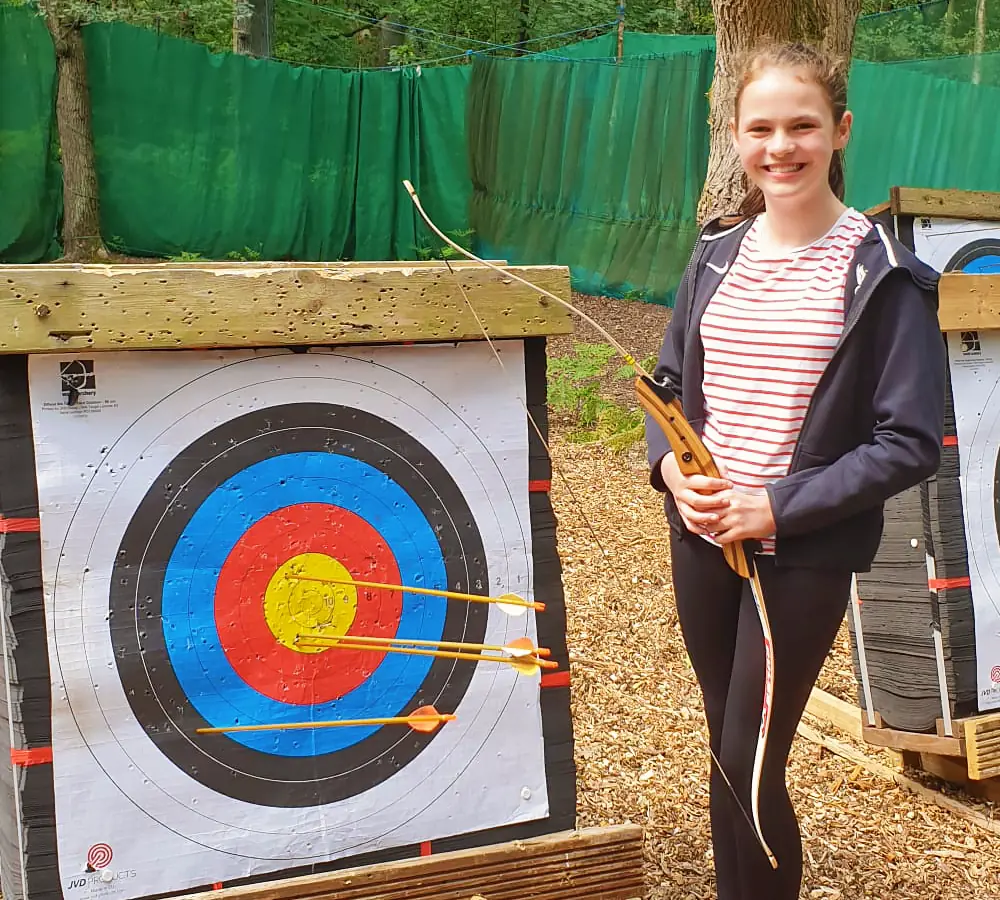 Smiling girl holding an archery bow stood next to an archery target with arrows in it