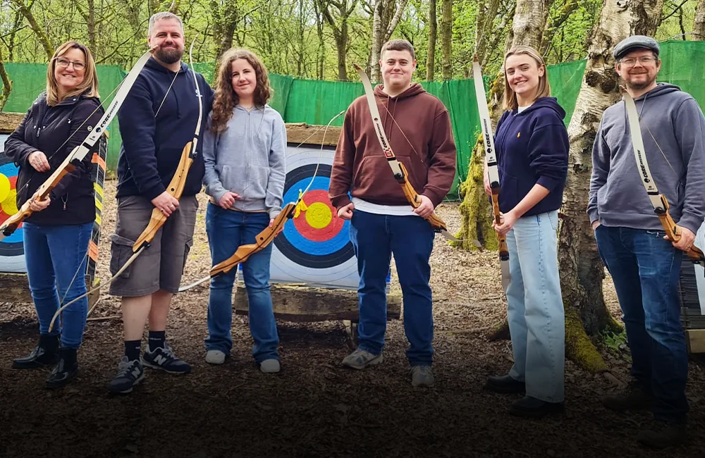 Six smiling people with archery bows stood on an outdoor shooting range