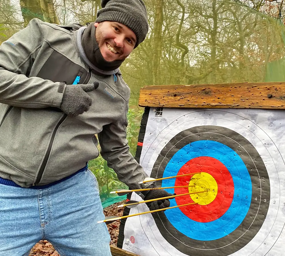 Grinning man giving a 'thumbs up' pointing at his arrows in the bullseye of an archery target