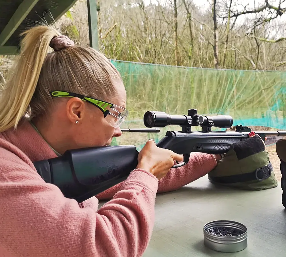 Lady wearing shooting glasses aiming an air rifle with a telescopic scope at Adventure Now Manchester