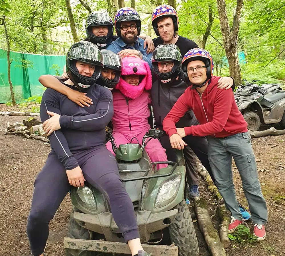 Group of guys stood together around a quad bike. Person in the middle wearing pink overalls