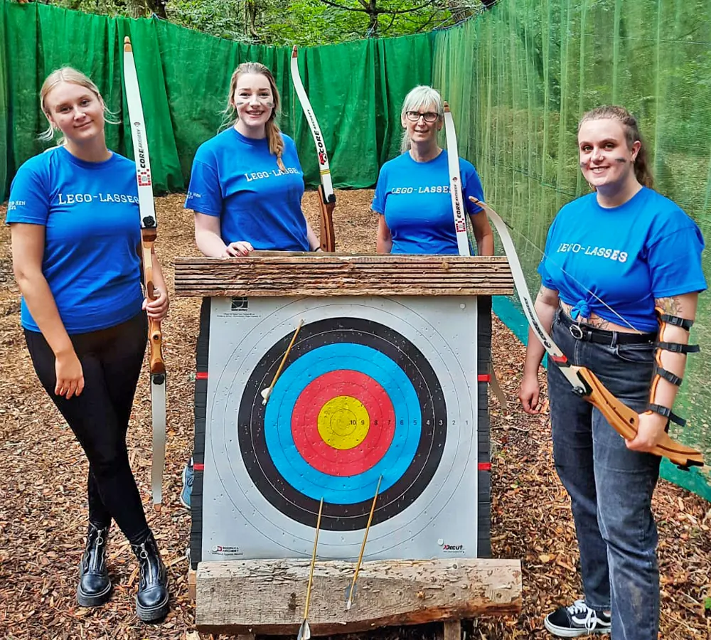 Four smiling women with matching shirts on a hen do, each holding a bow stood with an archery target