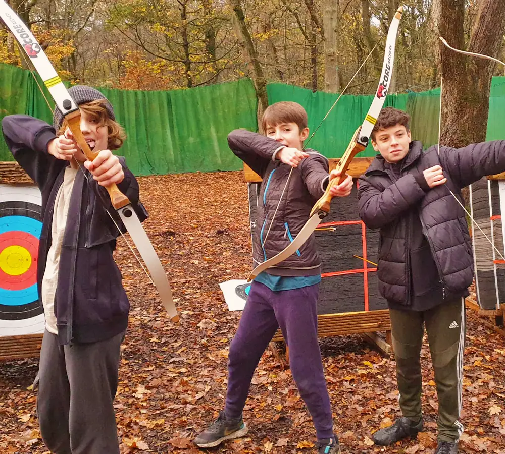 Three boys at a birthday party posing with archery bows in front of archery targets outdoors