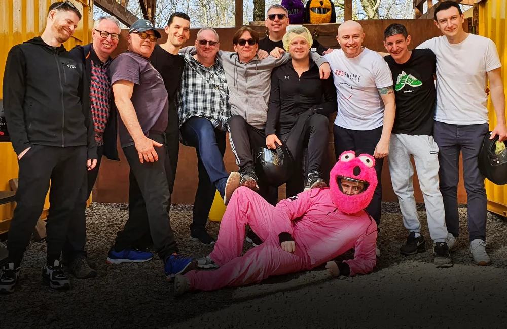 Large group of guys standing around the stag who is laying on the ground dressed in pink overalls and a pink crash helmet