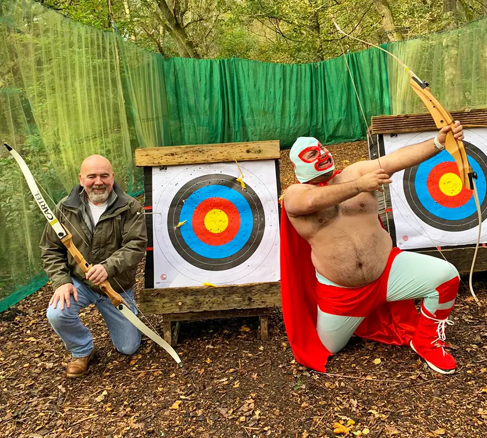 Two guys with archery bows posing in front of archery target. Man on the right is in fancy dress on a stag do