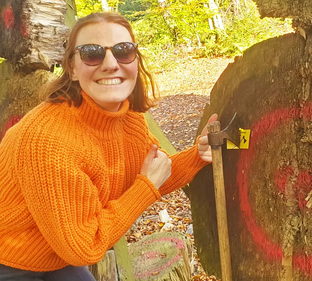 Smiling lady giving a 'thumbs up' stood with a throwing axe stuck in a large wooden target