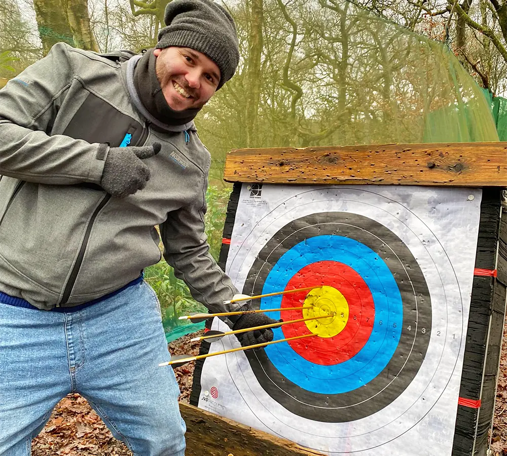 Grinning man giving a 'thumbs up' pointing at his arrows in the bullseye of an archery target