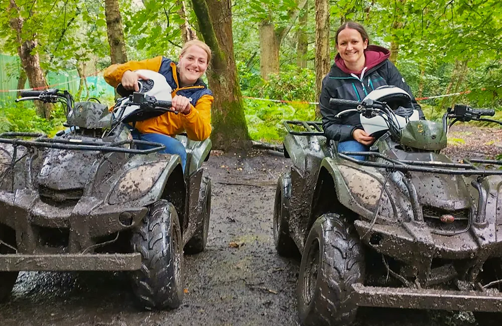 Two smiling ladies sat on muddy quad bikes holding crash helmets at Adventure Now Manchester