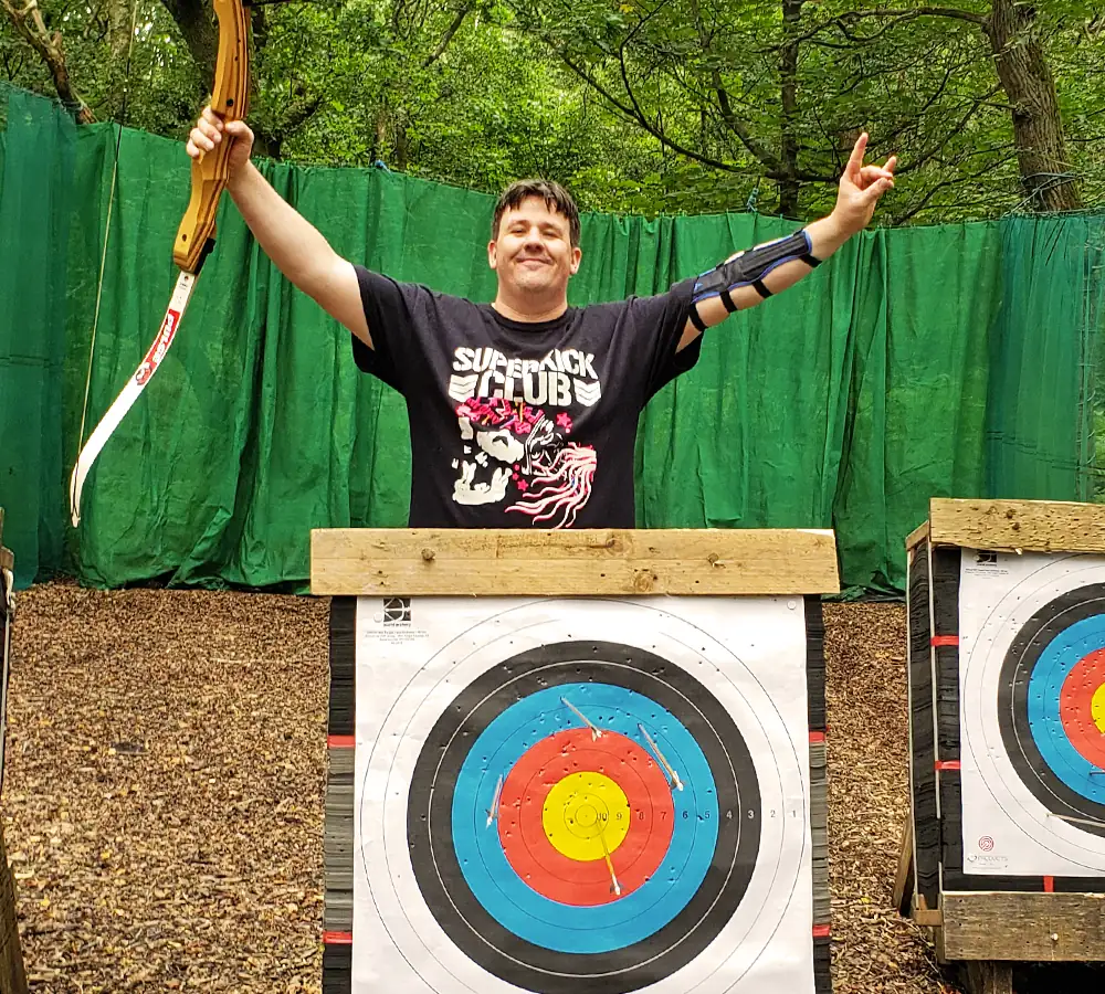 Happy smiling man holding an archery bow stood behind a target with several arrows in it